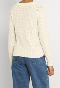 Krämfärgad, texturerad stickad tröja med rund halsringning och ribbad nederdel. Kombinerad med lösa blå denimjeans, vilket visar en avslappnad stil.