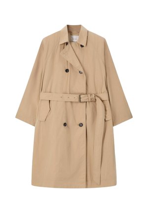 Frau steht mit kurzen braunen Haaren, trägt einen beigen Trenchcoat, ein weißes Oberteil, hellblaue Jeans, pinke Schuhe und hält eine große braune Handtasche.