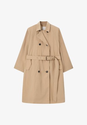 Vrouw staand met kort bruin haar, draagt een beige trenchcoat, wit shirt, lichtblauwe spijkerbroek, roze schoenen en houdt een grote bruine handtas vast.