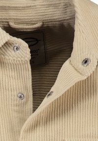 Colletto di camicia in corduroy beige con bottoni a pressione argentati. Strisce verticali testurizzate visibili sul tessuto. Etichetta visibile all'interno del colletto.