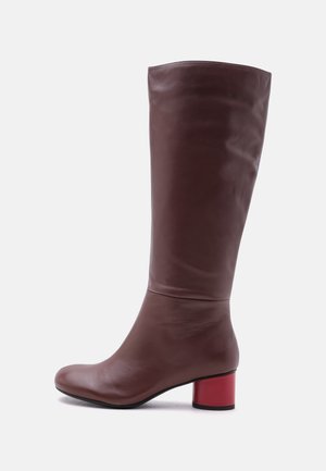 Botte haute en cuir marron avec un bout arrondi et un petit talon bloc rouge sur un fond clair uni.