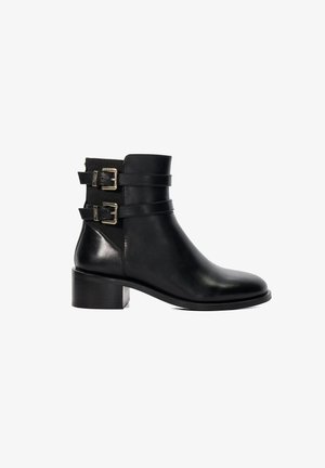 Bottine en cuir noir avec deux boucles décoratives, un bout rond et un talon bloc. Texture lisse avec une finition mate.