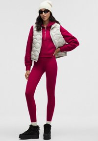 Kostium z różową bluzą z kapturem i legginsami, zestawiony z kremową pikowaną kamizelką. Dodatki obejmują białą czapkę i czarne wiązane botki.