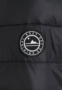 Svart vadderad jacka med en rund, broderad patch som har en vit design av ett berg och en sol, samt texten "ROXY EST 90" i en cirkulär layout.