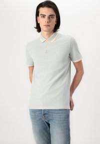 Polaire vert clair avec un col pêche, trois boutons et un contraste sur les manches. Porté avec un jean bleu clair.