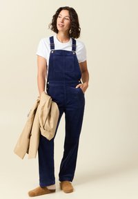 Navy-Walzstrampler aus Cord mit verstellbaren Trägern, Fronttaschen und Messingbeschlägen, kombiniert mit einem weißen T-Shirt und hellbraunen Schuhen.