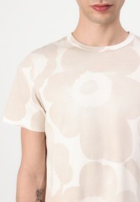 Korte ermer beige bomull T-skjorte med store lyse blomster mønstre og en liten hvit logo på brystet. Myk tekstur og avslappet passform.