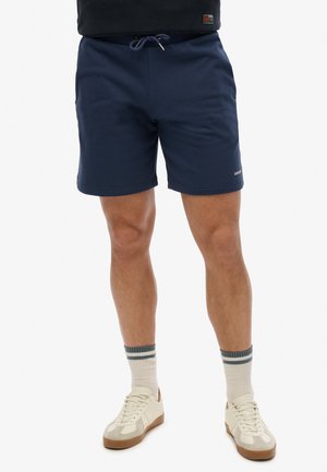 Person iført marineblå shorts med snørre, cremefarvede sneakers og cremefarvede sokker med grå striber, stående mod en hvid baggrund.