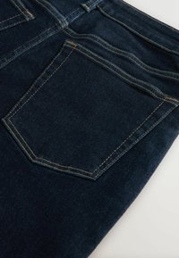 Jean en denim bleu foncé montrant la poche arrière avec une couture jaune contrastante sur un fond clair uni.