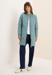 Giacca trapuntata di un azzurro chiaro con zip frontale, polsini a costine e design di lunghezza media. Abbinata a jeans scuri e sneaker bianche.