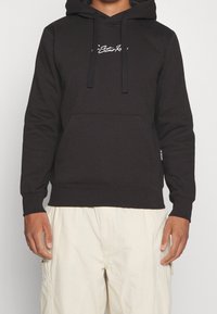 Felpa nera in misto cotone, con tasca frontale, cappuccio regolabile con cordino e logo bianco stampato sul petto. Tessuto liscio e vestibilità comoda.