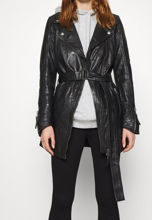 Osoba ubrana w czarną skórzaną kurtkę z paskiem, nałożoną na jasnoszarą bluzę z kapturem i czarne spodnie, stojąca na tle jednolitej białej ściany.