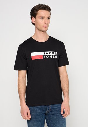 Mann trägt ein schwarzes T-Shirt mit weißem und rotem rechteckigem Logo mit der Aufschrift „JACK & JONES“, kombiniert mit blauer Jeans, steht vor einem einfarbigen weißen Hintergrund.