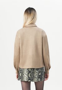 Beige textured jacket with a collared neck and buttoned cuffs, worn over a short snake-print skirt. Background is plain white.