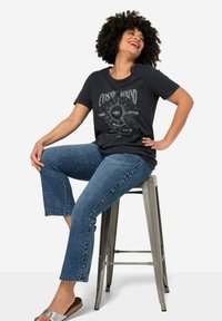 Camiseta de algodón negra con gráfico "COSMIC SOUND", corte relajado. El modelo lleva puesta unos pantalones vaqueros cortos de color azul claro y sandalias deslizantes plateadas, sentado en un taburete.