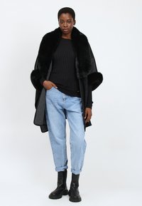 Donna che indossa un cappotto nero con bordo di pelliccia, un maglione nero a coste, jeans azzurri con risvolti e stivali neri alla caviglia, in piedi contro uno sfondo bianco.