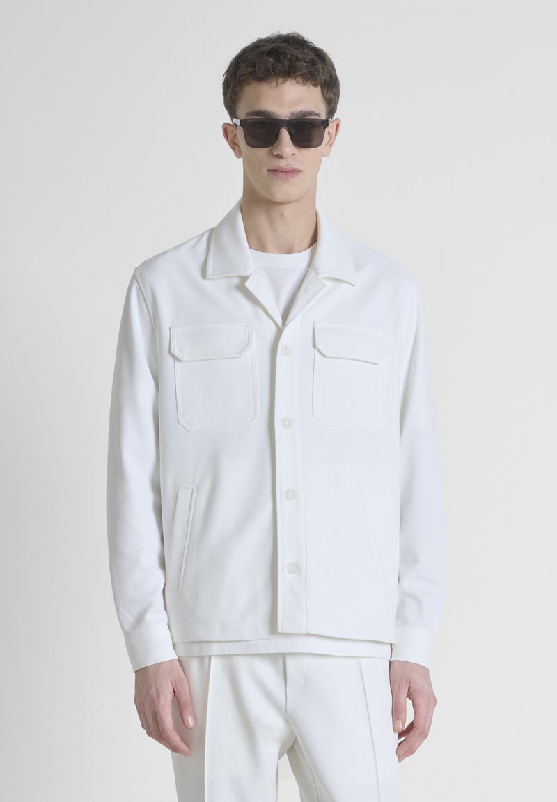 Chaqueta blanca de mezcla de algodón con dos bolsillos en el pecho, botones delanteros y cuello; llevada sobre una camiseta blanca sencilla.