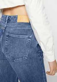 Jeans de ganga azuis com um bolso traseiro e um retalho de couro castanho claro rotulado "JDY". O tecido tem uma textura ligeiramente desbotada.