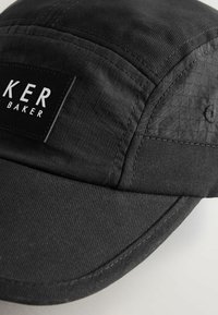 Mustast kangast nokamüts õmmeldud paneelidega ja esiküljel ristkülikukujuline plaastriga, kus on kiri "KER BAKER".