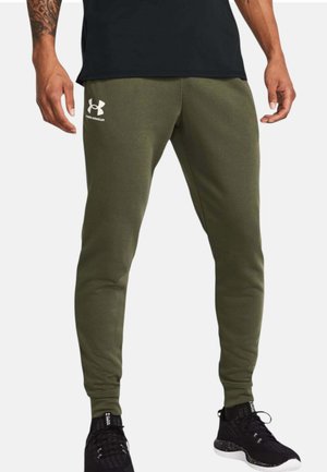Uomo che indossa pantaloni della tuta verde oliva Under Armour, sneakers nere e una maglia nera, in piedi con le braccia rilassate ai lati.