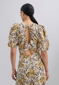 Blusa con estampado floral, mangas abullonadas, detalle de espalda descubierta, cierre con cordones y una vibrante combinación de colores amarillo, púrpura y negro sobre una tela clara.