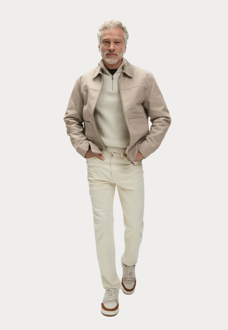 Uomo con capelli grigi che indossa una giacca beige, un maglione chiaro, pantaloni crema e sneakers beige mentre cammina con le mani in tasca.