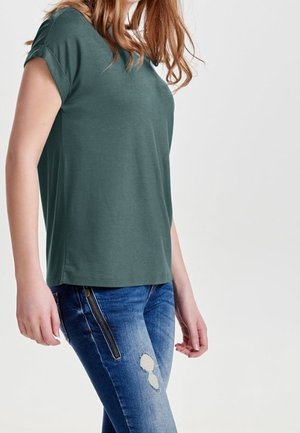 Persoon die een groen T-shirt met korte mouwen en blauwe versleten spijkerbroek met een ritssluiting draagt, staand tegen een effen achtergrond.