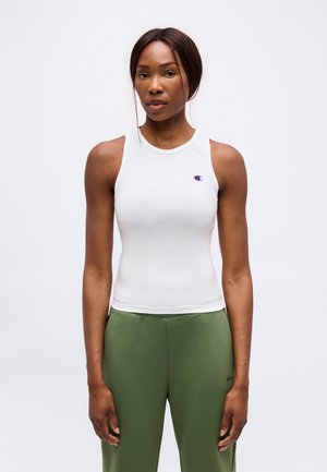 Donna con top sportivo bianco senza maniche e pantaloni verde oliva, in piedi rivolta in avanti su uno sfondo chiaro e uniforme.