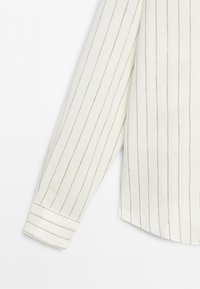 Chemise blanche à manches longues avec fines rayures grises verticales et un poignet boutonné sur un fond uni.