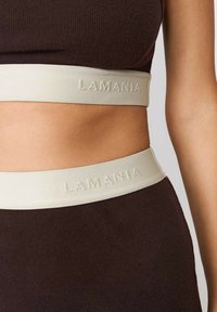 Hnedý rebrovaný crop top s svetlým béžovým elastickým pásom s prelisovaným logom "LAMANIA". Zamerajte sa na textúru a kontrast farieb.