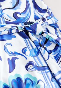 Tissu blanc avec des motifs floraux et tourbillonnants bleus noué en un nœud, montrant une texture lisse et un design vibrant de style aquarelle.