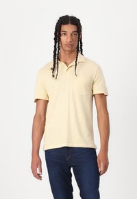 Lys gul piké med myk tekstur, kortermet og en brystlomme, kombinert med mørkeblå jeans. Enkelt design og avslappet passform.