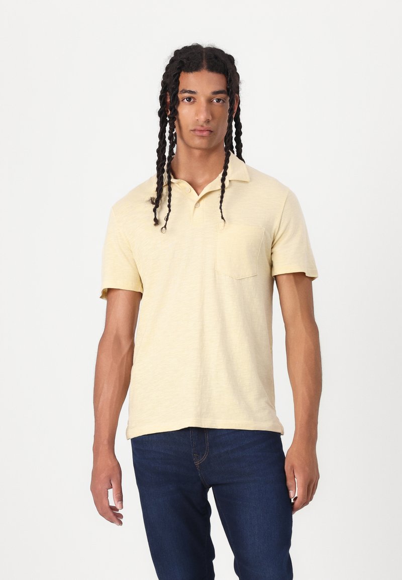Lys gul piké med myk tekstur, kortermet og en brystlomme, kombinert med mørkeblå jeans. Enkelt design og avslappet passform.
