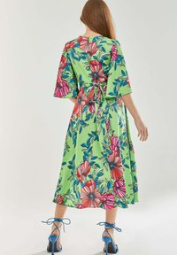 Floral-patterned, knee-length dress in green fabric, with bell sleeves, a tie waist, and vibrant pink and blue flowers. Paired with blue heels.