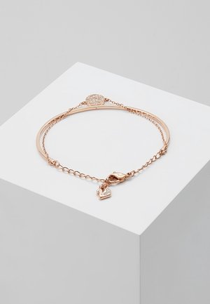 Hieno ruusukultainen rannekoru, jossa on ohut kaareva tanko, ketjulenkkirakenne, pieni papukaijalukko ja sydämenmuotoinen riipus, jossa on upotetut kristallit.