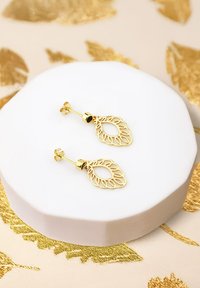 Orecchini a forma di foglia d'oro con ritagli intricati, montati su un semplice perno d'oro. Esibiti su un pedestal rotondo bianco contro uno sfondo strutturato.