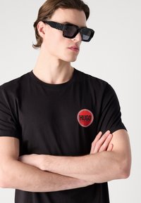 Black cotton T-shirt with round red logo patch featuring "HUGO" and checkered border; model wearing angular black sunglasses.