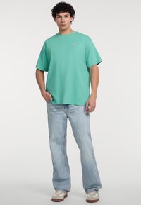 Mintgrünes T-Shirt mit kurzen Ärmeln aus Baumwolle, versehen mit einem kleinen dreieckigen Logo auf der Brust, kombiniert mit hellblauen Jeans und Sneakers.