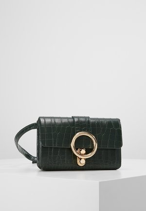 Donkergroene clutch van krokodillenleer met textuur, voorzien van een brede band en een grote ronde sluiting van goudkleurig metaal, geplaatst op een witte ondergrond.