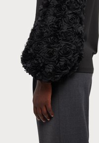 Sac à main noir avec un tissu texturé orné de roses noires en trois dimensions. Le sac a une forme arrondie et une apparence douce.