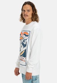 Witte sweatshirt met een grote grafische print van een sneaker met blauwe accenten en gestileerde tekst. Textuurstof en ribbels aan de mouwen als afwerking.