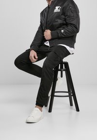 Giacca bomber nera con un logo sul petto, abbinata a pantaloni cargo neri e sneakers bianche, seduto su uno sgabello nero contro uno sfondo chiaro.