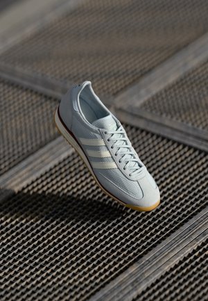 Lichtgrijze Adidas sneaker met witte strepen en rubberen zool die rust op een getextureerd metalen roosteroppervlak.