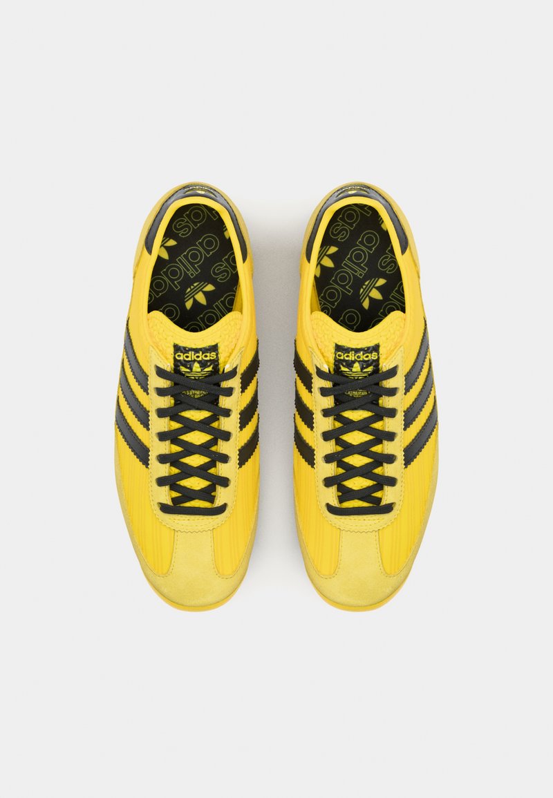 Zapatillas deportivas amarillas con franjas negras, parte superior texturizada, puntera redonda y cordones negros. Presenta un logotipo estampado en la lengüeta.
