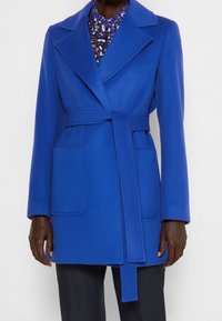 Abrigo de lana azul con un amplio collar, cintura ceñida y dos bolsillos de parche en la parte delantera. Textura suave y corte entallado. Camisa floral visible debajo.