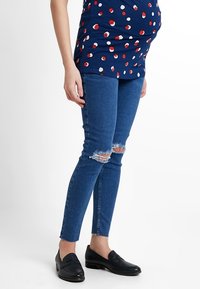 Zwangere persoon in een blauw met witte stippen top, gescheurde blauwe skinny jeans en zwarte instappers, staand voor een plain witte achtergrond.