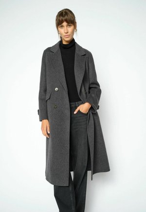 Donna con un lungo cappotto doppiopetto grigio scuro su un dolcevita nero e pantaloni ampi grigio scuro. Ha una mano in tasca.