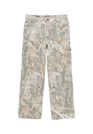 Personne portant un pantalon camouflage à jambes larges et des baskets marron foncé, debout devant un fond uni, les bras croisés.
