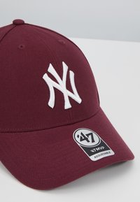 Burgunder rød baseballcaps laget av stoff, med en hvit brodert "NY"-logo og en label som indikerer at den er justerbar.