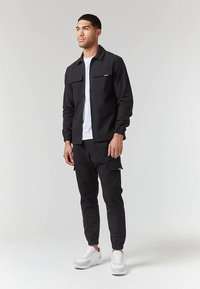 Black zip-up jacket with two chest pockets, paired with black cargo pants. White t-shirt underneath, and white sneakers. Smooth fabric texture.
